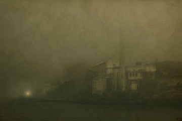 A moody, sepia-toned photograph of an abandoned industrial building on Alcatraz Island in San Francisco, shrouded in heavy fo